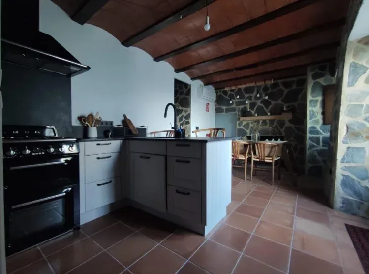 Imagen de La cocina se encuentra en la planta baja a pié del patín de la casa.La cocina mide 16m2 y tiene chimenea y wifi. Tiene luz natural que le da la ventana y la puerta principal de la casa. El techo es de bovedillas que junto a las paredes de piedra le da un carácter único.La cocina está equipada con horno, nevera, microondas, tostadora, hervidor eléctrico, cafetera Dolce Gusto, sandwichera, batidora y todos los utensilios necesarios para cocinar y tomarse algo (cápsulas café, tés e infusiones, azúcar, sal, pimienta, aceite). Existen dos zonas habilitadas para comer, una en la barra de la cocina y otra en la mesa que se encuentra delante de la chimenea.Disponemos de trona.