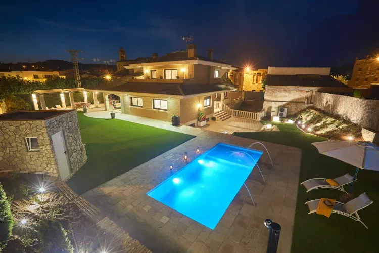 Imagen de Vista del Jardín, se puede observar de derecha a izquierda. Muro con decoración de manta de coco y diferentes arbustos de la zona compuesta entre piedras de la época. Zona césped y piscina realizada con mármol travertino. Zona intermedia de césped y vista de la Carpa con el toldo eléctrico.